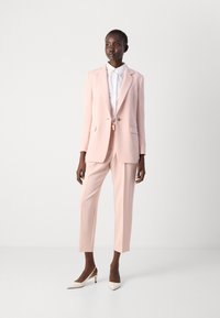 Completo su misura rosa chiaro con blazer a revers e pantaloni affusolati, abbinato a una camicia bianca fresca e décolleté beige a punta.