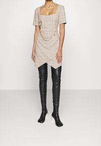 Beżowa sukienka w kratę z krótkimi rękawami, kwadratowym dekoltem, asymetrycznym dołem i łańcuszkiem w talii, zestawiona z czarnymi skórzanymi wysokimi botkami.