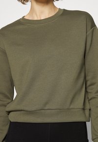 Olive green sweatshirt in a soft, smooth fabric with a crew neck and ribbed hem. Features long sleeves and relaxed fit, paired with black pants.