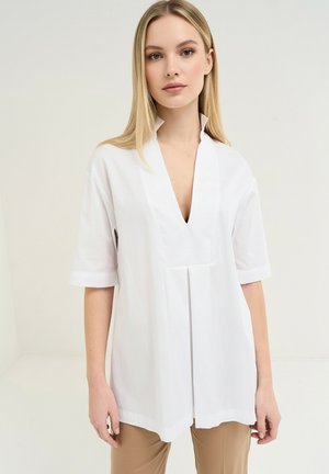 Femme blonde portant un chemisier blanc ample à manches courtes avec un profond col en V et un pantalon beige, debout devant un fond uni.