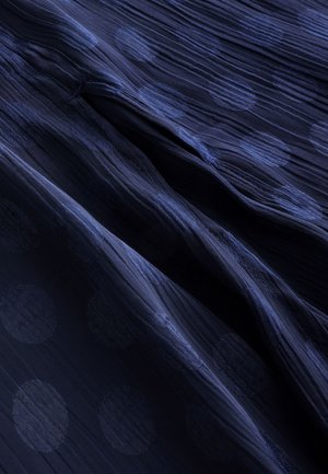 Tamnoplava tkanina s teksturiranim uzorkom istaknutih kružnih točkica. Materijal ima mekan, tečan izgled i lagani završni sloj.