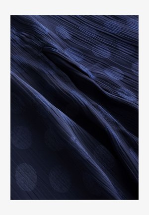 Tamnoplava tkanina s teksturiranim uzorkom istaknutih kružnih točkica. Materijal ima mekan, tečan izgled i lagani završni sloj.