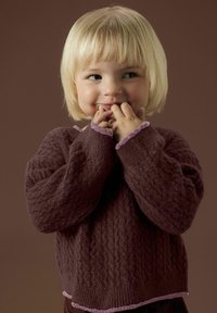 Pull en maille marron avec un motif texturé, doté d'une bordure lilas à volants aux poignets et à l'ourlet. Modèle souriant, mains près du visage.