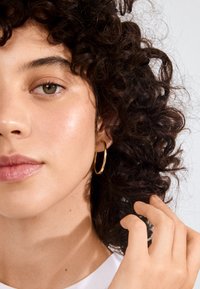 Orecchino a cerchio dorato all'orecchio di una donna, corto e circolare con una finitura liscia. Capelli ricci castani e tonalità della pelle chiara. Maglietta bianca semplice.