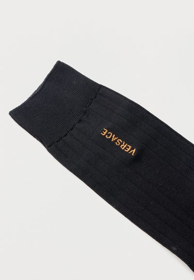 Μαύρη ριμπ καλτσοδέτα με κεντητό λογότυπο «VERSACE» σε χρυσή ραφή στη πλευρά κοντά στον αστράγαλο.