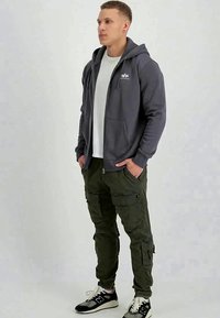Jovem a vestir um hoodie cinza escuro com fecho, uma t-shirt branca, calças cargo verdes e ténis preto e branco, posicionado frente a um fundo claro.