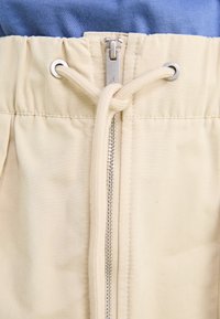 Pantaloni bej cu talie elastică, având un fermoar argintiu și șnur, și un top albastru parțial vizibil deasupra.