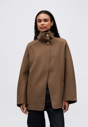 Vrouw met krullend haar, gekleed in een bruine wollen jas met hoge kraag en twee gouden knopen, zwarte broek, staande tegen een effen lichte achtergrond.