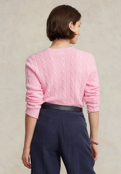 Femme aux cheveux courts et bruns portant un pull en maille torsadée rose et un pantalon bleu marine avec une ceinture noire, vue de dos sur un fond beige.