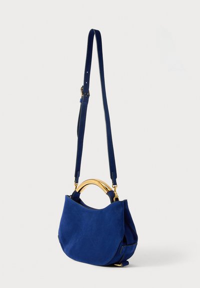 Malá tmavě modrá semišová kabelka přes rameno s oblým zlatým kovovým držadlem a nastavitelným popruhem na bílém pozadí.
