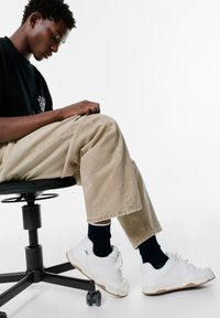 Białe sportowe sneakersy z teksturowanym wzorem, połączone z ciemnymi skarpetkami i beżowymi spodniami o szerokich nogawkach. Siedząc na czarnym ergonomicznym krześle.