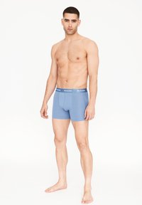 Světle modré trenýrky s těsným střihem, měkkou látkou a kontrastním páskem s bílým nápisem. Model stojí bos a v neutrální pozici.