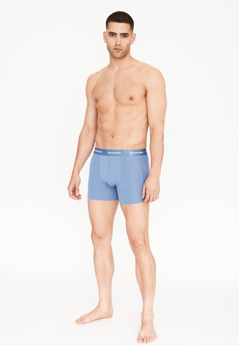 Světle modré trenýrky s těsným střihem, měkkou látkou a kontrastním páskem s bílým nápisem. Model stojí bos a v neutrální pozici.