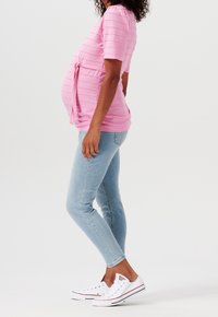 Růžový pruhovaný těhotenský top se zavazováním v pase, doplněný světle modrými úzkými džíny a bílými teniskami. Materiál je měkký s texturovaným vzorem.