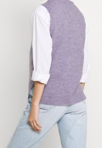 Světlofialová pletená vesta s kulatým výstřihem, nošená přes bílou košili s knoflíky, doplněná světle modrými džínami.