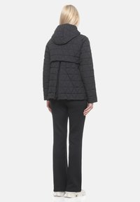 Schwarze, gesteppte Jacke mit Kapuze, die ein dreieckiges Muster und einen verlängerten Saum hinten aufweist, kombiniert mit schwarzen Schlaghosen und hellen Sneakers.