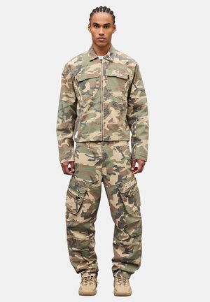 Männliches Model trägt eine grün-beige Tarnjacke und Cargohosen mit sandfarbenen Stiefeln, steht vor einem schlichten hellen Hintergrund.