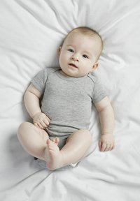 Un grenouillère en coton gris sur un bébé allongé sur un drap blanc, avec des manches courtes et un col arrondi, montrant une peau douce et de petits pieds.