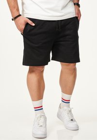 Schwarze Baumwollshorts mit elastischem Bund, seitlichen Taschen und lockerer Passform. Kombiniert mit weißen Sneakers und mehrfarbigen gestreiften Socken.