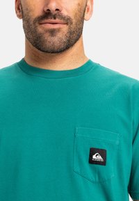 Tyrkysové bavlněné tričko s kulatým výstřihem, krátkými rukávy a náprsní kapsou na levé straně, která je ozdobena černou nášivkou loga.