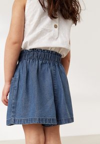 Jeansskørt med elastisk talje og sidelommer. Blå farve med afslappet pasform og frynset kant. Hvid top delvist synlig.