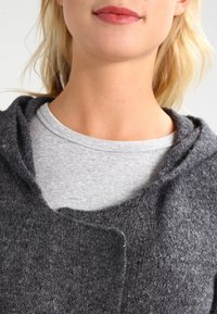 Szara filcowa odzież z kapturem o miękkiej teksturze, z zaokrąglonym dekoltem. Nosi się ją na jasnoszarą, dopasowaną koszulkę.