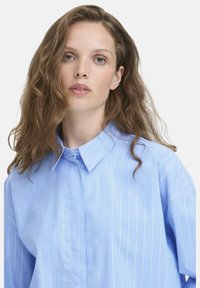Light blue button-up shirt with thin white vertical stripes, featuring a pointed collar and relaxed fit with short sleeves.