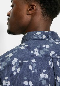 Blumenhemd aus dunkelblauem Denim-Stoff, mit weißen Blumen und grünen Stängeln. Enthält ein Knopfdetail am Kragen.