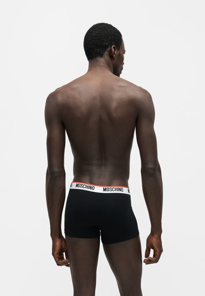 Άνδρας μοντέλο φαίνεται από πίσω φορώντας μαύρα μποξεράκια Moschino με λευκή μέση και μαύρο λογότυπο, στέκεται με φόντο απλό λευκό.