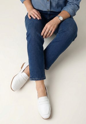 Person iført blå jeans, en denimskjorte, et armbåndsur, ringe og hvide lædersko uden snørre, siddende med benene krydsede.