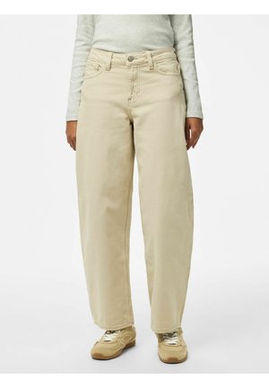 Person iført beige vidde bukser med lommer, beige sneakers og en lysegrå langærmet top, stående mod en hvid baggrund.