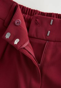 Burgundské kalhoty s hladkou texturou, elastickým pasem, zipovým zapínáním a kovovými sponami s knoflíkovým detailem.