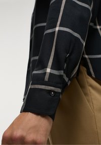 Chemise à manches longues à carreaux noirs avec un motif à carreaux gris clair contrastant, des poignets à boutons et une texture de tissu douce avec une légère brillance.