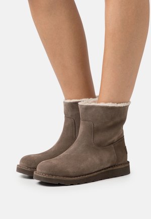 Bottines en daim marron avec doublure en polaire blanche portées sur des jambes nues, présentées sur un fond blanc.