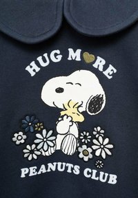 Námornícky sveter má grafiku Snoopyho, ktorý drží žltého vtáka, obklopeného farebnými kvetmi. Text znie: "OBJÍMAJ VIAC PEANUTS KLUB."