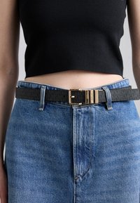 Čierny kožený opasok so zlatou kovovou sponou, nosený cez priliehavý čierny crop top a vysoko pásové modré džínsy. Viditeľný štruktúrovaný povrch.