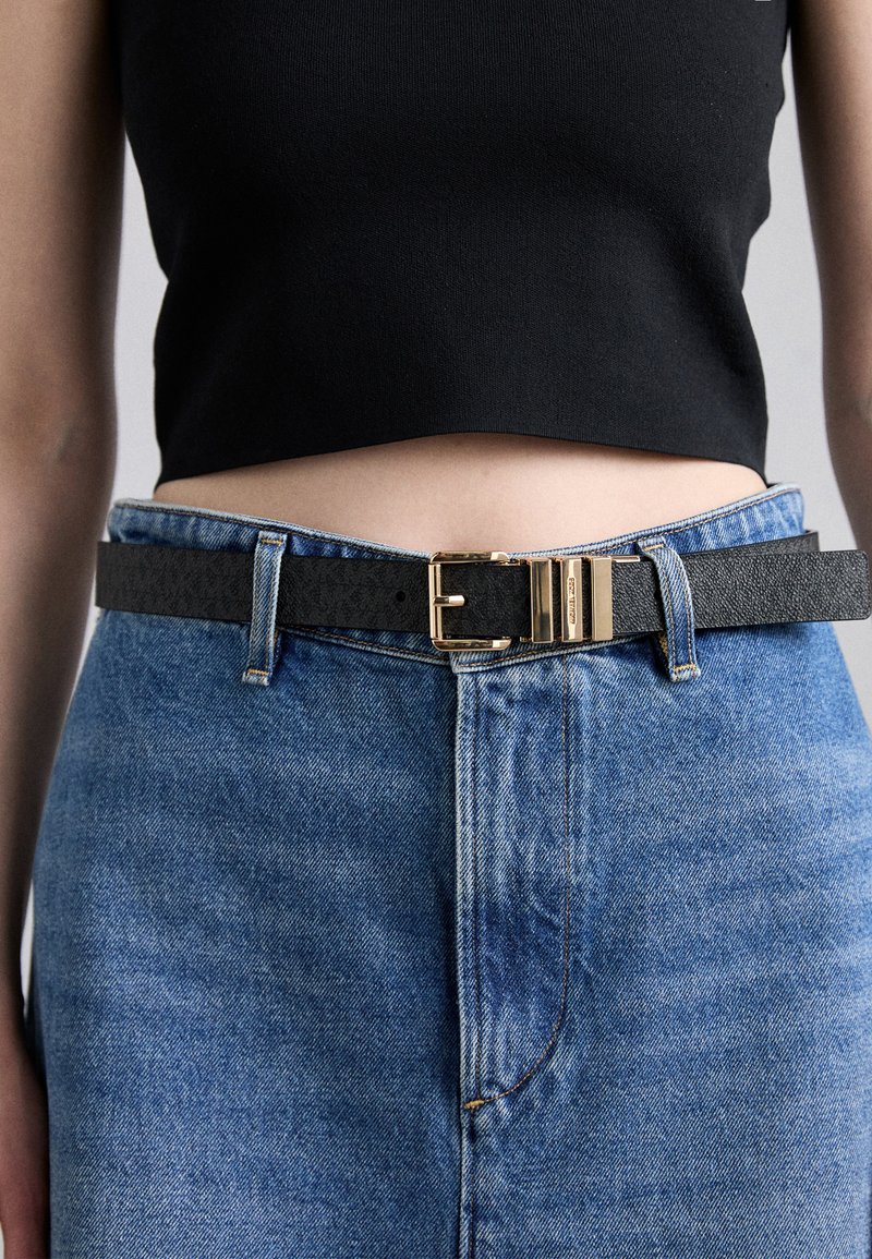 Čierny kožený opasok so zlatou kovovou sponou, nosený cez priliehavý čierny crop top a vysoko pásové modré džínsy. Viditeľný štruktúrovaný povrch.