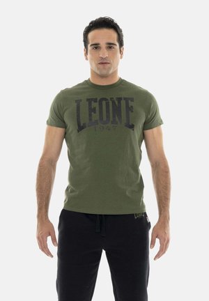 Man staat recht vooruit kijkend, met een olijfgroen "LEONE 1947" T-shirt en een zwarte broek tegen een effen lichte achtergrond.