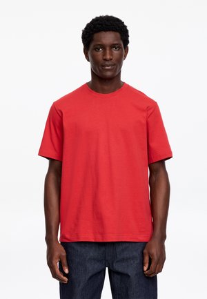 Uomo in piedi rivolto verso lo spettatore con una t-shirt rossa semplice a maniche corte e jeans blu scuro su sfondo bianco.