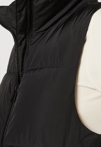 Svart vadderad puffväst med hög krage, med dragkedjestängning och strukturerad tyg, bärs över en ljusfärgad topp.