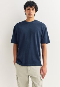 Granatowy bawełniany t-shirt o luźnym kroju, z krótkimi rękawami, okrągłym dekoltem i subtelną fakturą. Noszony z jasnobeżowymi spodniami typu cargo.