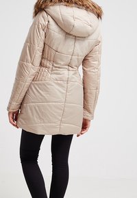 Giacca a vento beige con cappuccio, caratterizzata da un bordo in pelliccia ecologica, texture trapuntata e silhouette aderente. La vista posteriore mostra dettagli delle cuciture verticali.