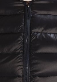 Veste matelassée noire avec des sections cousues horizontalement, texture lisse et fermeture éclair intégrale avec une tirette en métal argenté.