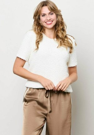 Lachende vrouw met lang blond haar, gekleed in een wit gestructureerd shirt met korte mouwen en losse beige satijnen broek, tegen een effen achtergrond.