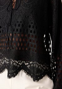 Svart blondebluse med en gjennomsiktig design, med et teksturert mønster av åpne diamanter, intrikat broderi og en bølget kant.