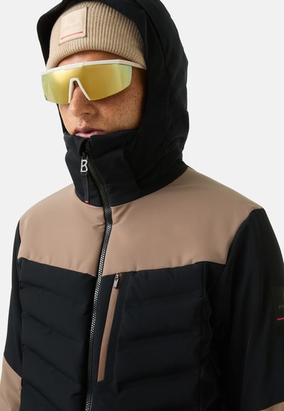 Schwarz-beige isolierte Jacke mit hohem Kragen, Reißverschluss auf der Vorderseite und Brusttasche. Begleitet von Sonnenbrille und beigem Beanie.