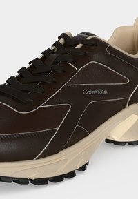 Σκούρο καφέ αθλητικό παπούτσι Calvin Klein με μαύρα κορδόνια, κρεμ σόλα και λευκές ραφές σε λευκό φόντο.