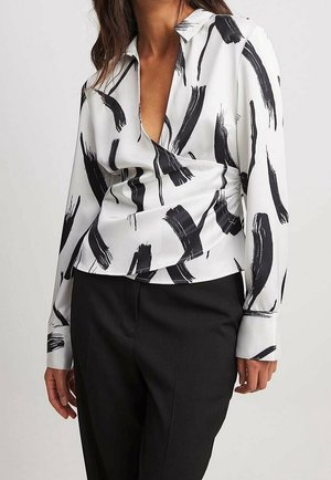 Kobieta ubrana w białą bluzkę z kopertowym dekoltem w czarne wzory przypominające pociągnięcia pędzla oraz czarne spodnie na jednolitym tle.