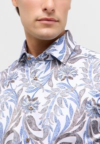 Witte shirt met blauw en bruin bloemmotief; heeft een spreidkraag, knoopsluiting aan de voorkant en een gladde, lichte stofstructuur.