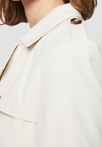 Světle béžová bunda s límcem, detaily na rameni a měkkou texturou. Látka vypadá hladce s upravenými švy.